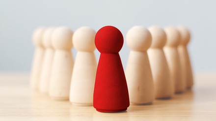 Nine small wooden pins on a wooden desk, in an arrow formation with the only red pin in the middle.