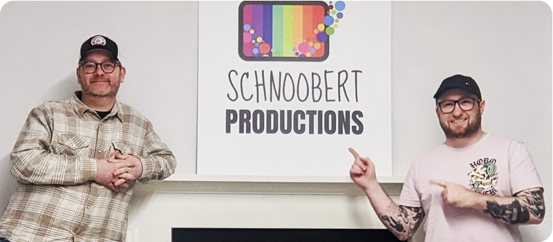 Two men dressed in casual attire and caps stand either side of a fireplace which has a board with the Schnoobert Productions logo on its mantlepiece.