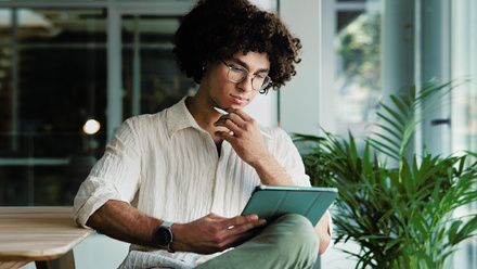 A person sitting in a light, glass office with plants, intently reading something on an iPad with a green cover.