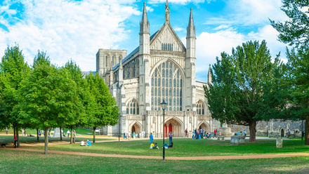 Summer time view of Winchester Cathedral west front in Hampshire surrounded by trees.
