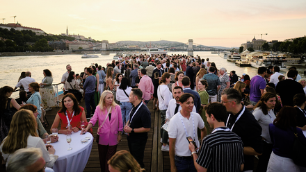 Photo of a large networking event happening on the upper deck of a boat with the Budapest skyline at sunset in the background.