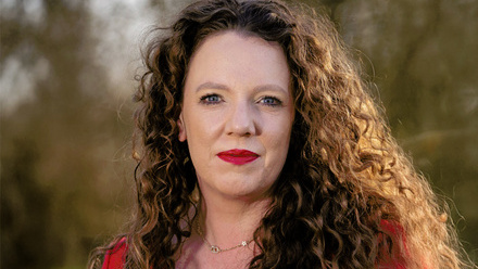 Headshot: Kate Beale, a woman with dark, curly hair wearing a red top and red lipstick.