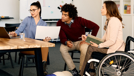 Photo of three people in a creative office environment, one is in a wheelchair holding a notebook. All three are looking toward a laptop on a desk.