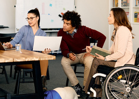 Photo of three people in a creative office environment, one is in a wheelchair holding a notebook. All three are looking toward a laptop on a desk.