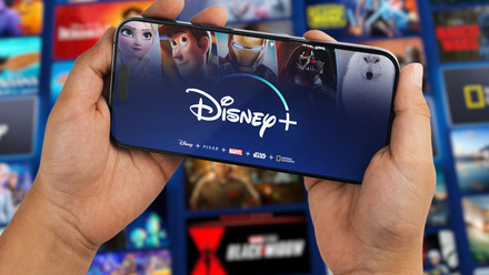 Two hands holding a smartphone with the Disney+ logo on the screen. In the background beyond is a wall of blurred images of Disney+ shows.