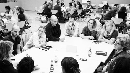 A room full of groups of people sat around large rectangle tables, they are engaged and listening attentively to each other.
