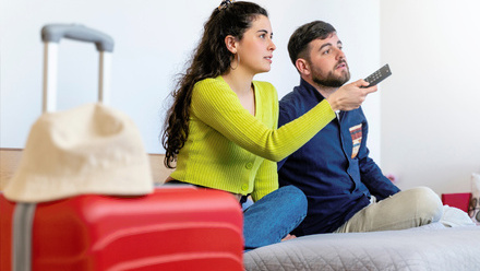 A couple sits on a bed in a hotel room, holding up a TV remote and both looking toward the screen out of shot.