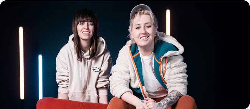 Two women pose in light hoodies in a dark room with white bar neon lights in the background.