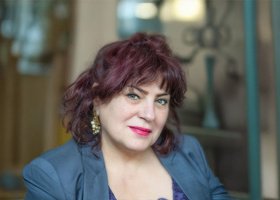 Headshot: Sepi Chakaveh, a woman with short purple hair wearing gold statement earrings and a grey blazer.