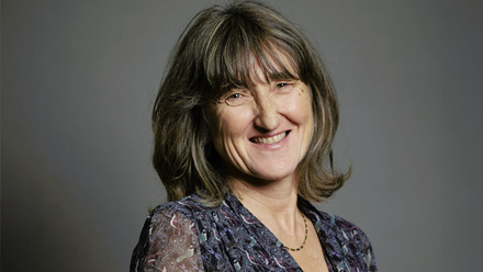 Headshot: Baroness Kidron, a woman with shoulder-length brown hair wearing a patterned, dark blue top and a gold necklace.