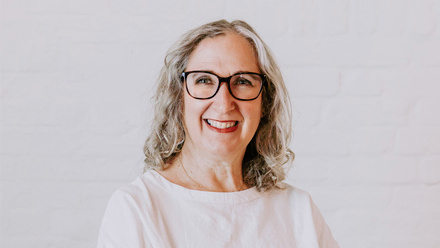Headshot: Mandy Hall, a woman with wavy grey hair wearing black glasses and a white top.