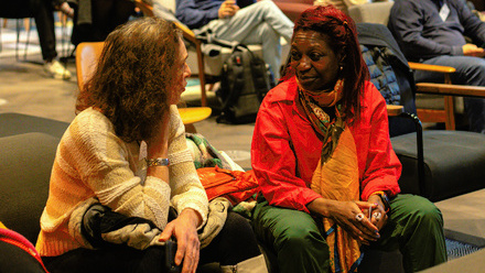 Photo of two women sat down talking. More people can be seen in the background, sat in chairs waiting for an event to begin.