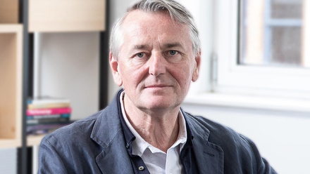 Headshot: Max Rumney, a man with short grey hair. He is wearing a dark blue suit jacket with a light shirt underneath.