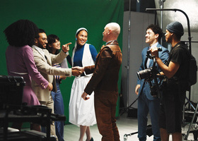 Group of people smiling and shaking hands on a film set with a green screen and camera equipment.