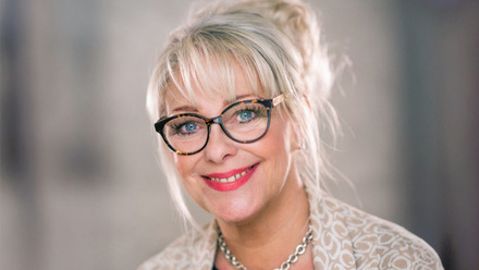 Headshot: Dawn McCarthy Simpson, a woman with blonde hair tied back in a bun and a fringe, wearing glasses, pink lipstick, a necklace and cardigan.