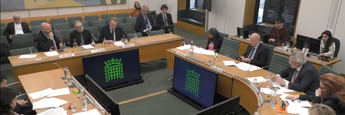 Image: Pact CEO John McVay giving evidence to MPs. A formal room with an oval desk which 6 MPs are sitting behind. In front of them, a long table where John McVay and two others sit facing the MPs.