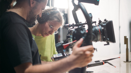 A director with glasses and headphones stands next to a camera operator holding a small cinema camera on a rig.