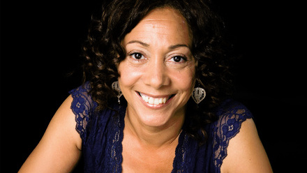 Headshot: Thomasina Gibson, a woman with dark brown, curly hair wearing statement earrings and a blue lace top.