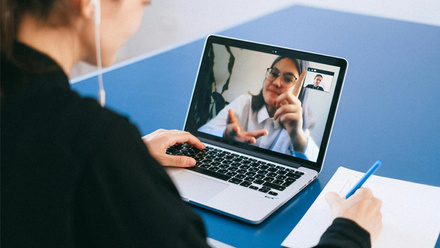 Person sat at a blue desk wearing wired headphones and writing on a pad of paper while talking on a virtual call to another person on a laptop.