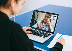 Person sat at a blue desk wearing wired headphones and writing on a pad of paper while talking on a virtual call to another person on a laptop.
