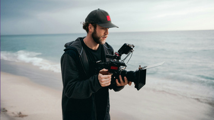 Camera operator holding a camera on a beach with the sea in the background.