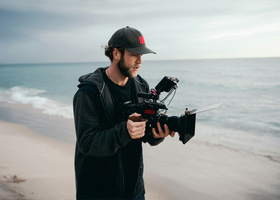 Camera operator holding a camera on a beach with the sea in the background.
