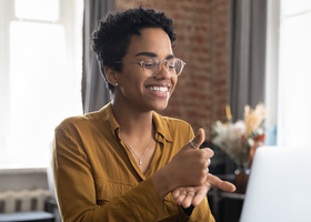 Photo of a woman sitting at a desk in front of a laptop. She is signing thumbs up.