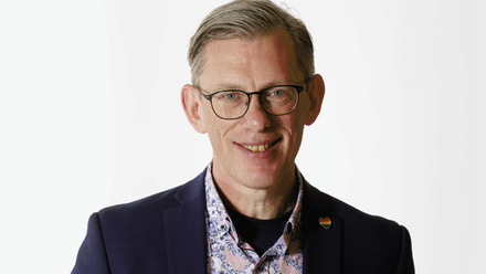 Headshot: Paul Byrne, a man with grey hair slicked to the side, wearing glasses, a patterned shirt, a blue blazer and an LGBTQ+ pin.