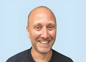 Headshot: Danny Fenton, a bald man with a stubble beard wearing a black t-shirt.