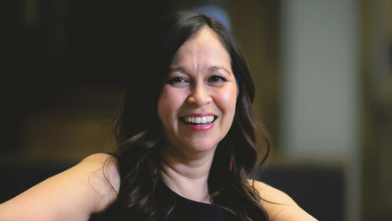 Headshot: Mel Rodrigues, a woman with dark brown wavy hair, wearing a black top.