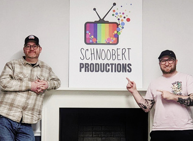 Two men dressed in casual attire and caps stand either side of a fireplace which has a board with the Schnoobert Productions logo on its mantlepiece.