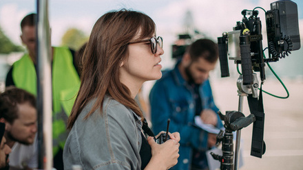 Photo of a woman with long brown hair on set, holding a walkie talkie and looking at a monitor. Crew members are working in the background.