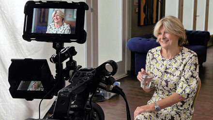 A woman wearing a summer dress and holding a glass of water sits on a chair in front of a large broadcast camera with her face seen on the monitor.