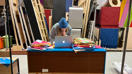 A person leaning over a desk on a laptop in a workshop surrounded by colourful materials and wooden frames.