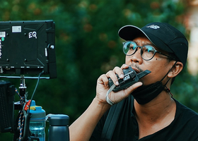 Photo of a director on location, he is standing behind a monitor and holding a walkie-talkie up to his mouth.