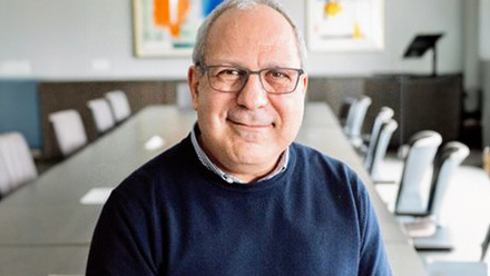 Headshot: Ed Hersh, a man with very short grey hair, wearing glasses and a navy blue jumper with a white shirt showing at the collar.