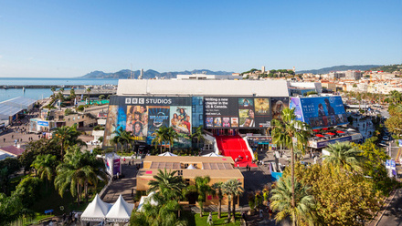 Panoramic view of MIPCOM Cannes featuring a blue sky, coastline, palm trees and a large building with a red carpet.
