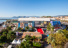 Panoramic view of MIPCOM Cannes featuring a blue sky, coastline, palm trees and a large building with a red carpet.