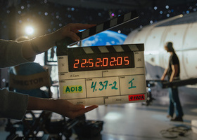 A crew member holds a digital clapperboard in front of a sci-fi set with production lights and a crew member in the background.