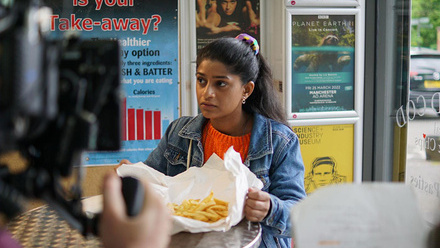 Actor sat at a table in a takeaway with a paper parcel of chips looking at a camera. There are framed posters on the wall in the background.