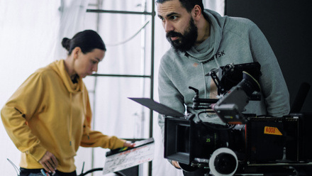 On-set, two crew members are in action, one standing over and adjusting a large cinema camera and the other walking with a clapper in hand.