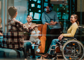 Photo of a group of office workers in a breakout room, including a woman in a wheelchair who is smiling at the person speaking.