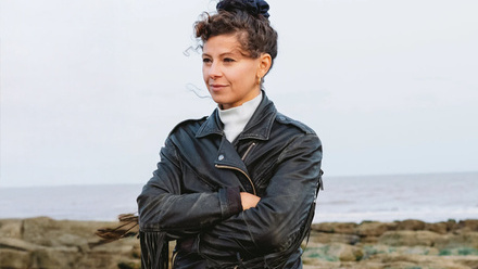 A person with brown curly hair tied up in a bun, wearing a white turtleneck and a black jacket, looking into the distance.