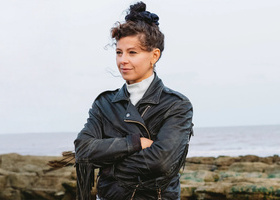A person with brown curly hair tied up in a bun, wearing a white turtleneck and a black jacket, looking into the distance.