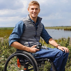 Photo: Steve Brown, a white man with blond hair is sitting in a wheelchair with a pair of binoculars in his hand. The background is a lush and green countryside setting.