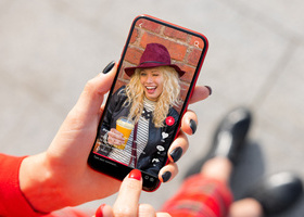 A phone held by a woman with red nail polish, the screen is open on a social media site showing a blonde-haired woman smiling and holding a drink.