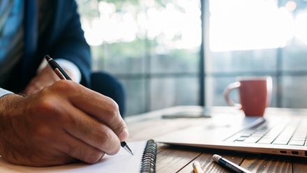 Photo close up of a hand writing on a notepad on a desk, with a mug and open laptop in the background.