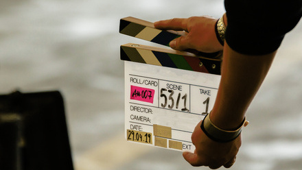 Photo close up of a clapper board with writing and coloured tape on it being held in front of a large camera with a street in the background.