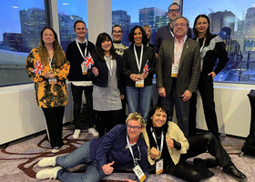 A group of 10 UK Indies delegates with Pact's Ross Lewis, posing for the camera with some mini-Union Jack flags in front of a large window.