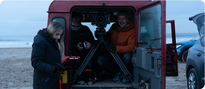 A small film crew works from the back of a red van parked on a beach, with the rear door open and a large camera on a tripod inside.